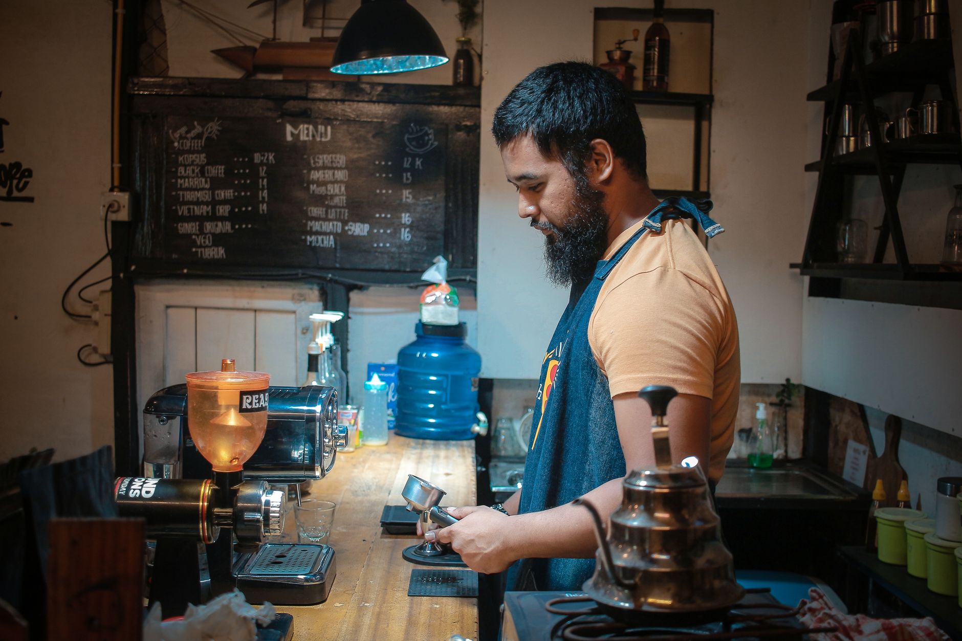 A barista with a beard preparing coffee in a cozy cafe with a chalkboard menu background.
